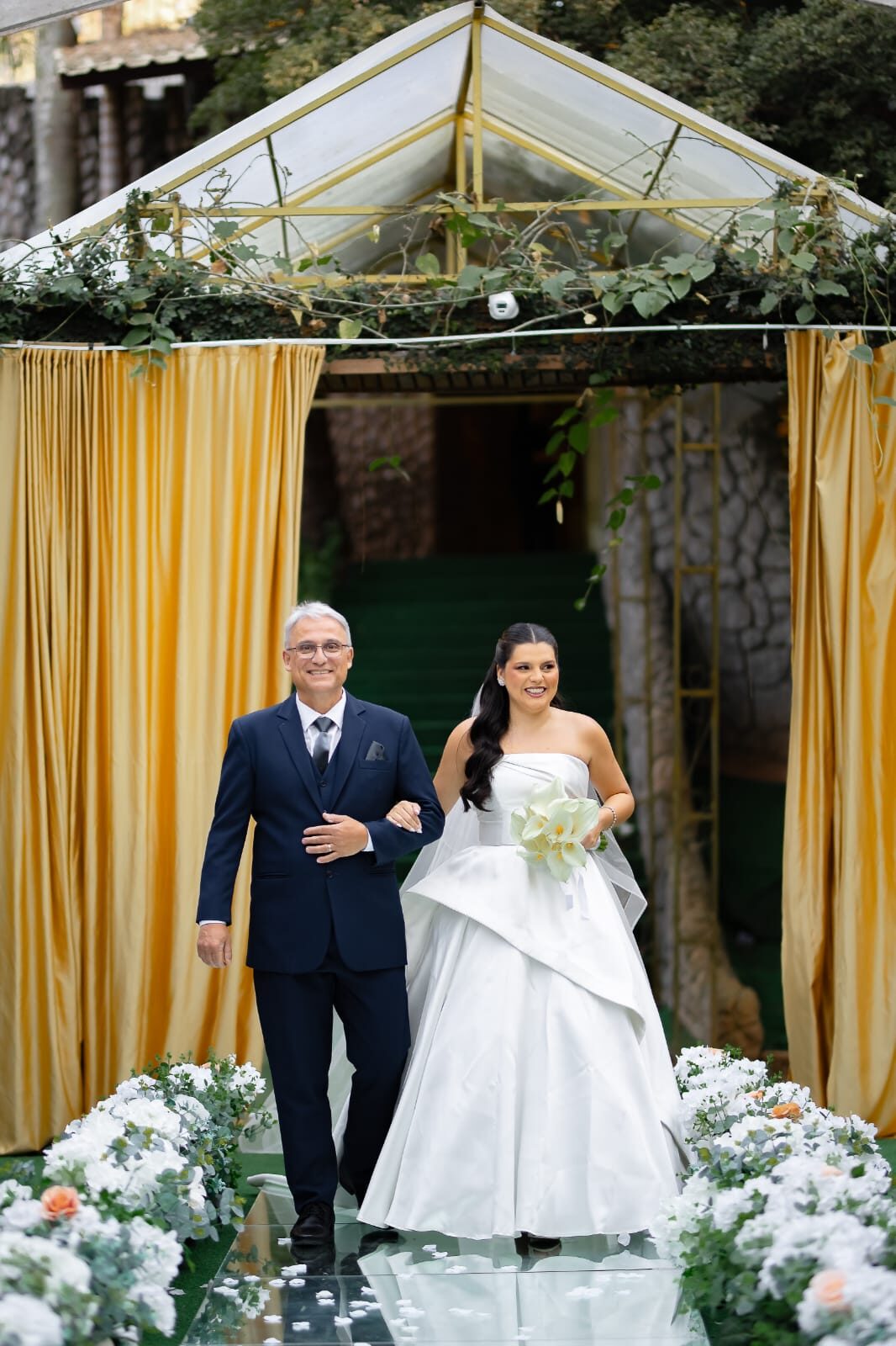 Noiva sendo conduzida pelo pai na entrada com cortinas douradas